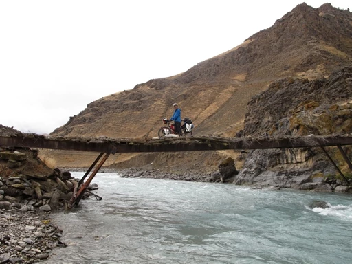 Radfahrer auf einer schmalen, alten Holzbrücke über einen türkisfarbenen Gebirgsfluss, umgeben von felsiger Berglandschaft