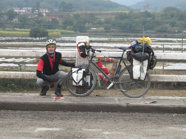 Fahrradtourist in rot-schwarzer Jacke sitzt neben vollbepacktem Reisefahrrad an Straßenböschung mit Berglandschaft im Hintergrund