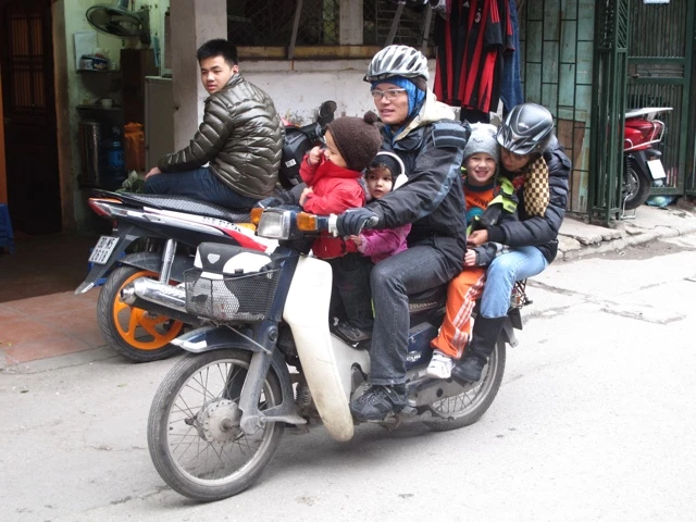 Mehrere Personen, darunter Kinder, dicht gedrängt auf einem kleinen Motorroller in einer städtischen Straßenszene