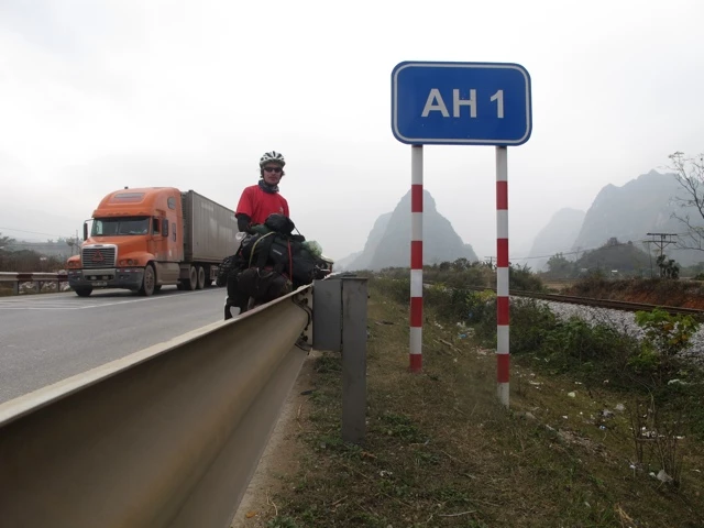 Ein Radfahrer in roter Jacke sitzt an einem Straßenrand der AH1-Fernstraße, im Hintergrund ein orangefarbener Lastwagen und nebelige Berge
