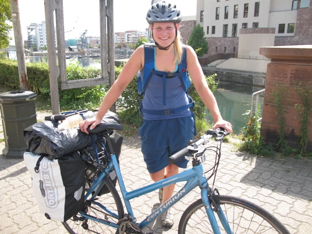 Junge Radfahrerin mit blauem Fahrrad, Helm und Packtaschen an urbaner Wasserstraße