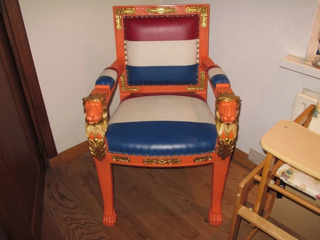 Historischer Sessel mit ornamentalen Verzierungen in Rot, Weiß und Blau, goldene Löwenkopf-Armlehnen, auf Holzboden