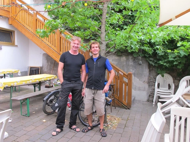 Zwei Männer in Freizeitkleidung stehen in einem Innenhof mit Holztreppe, Gartenmöbeln und Grünpflanzen