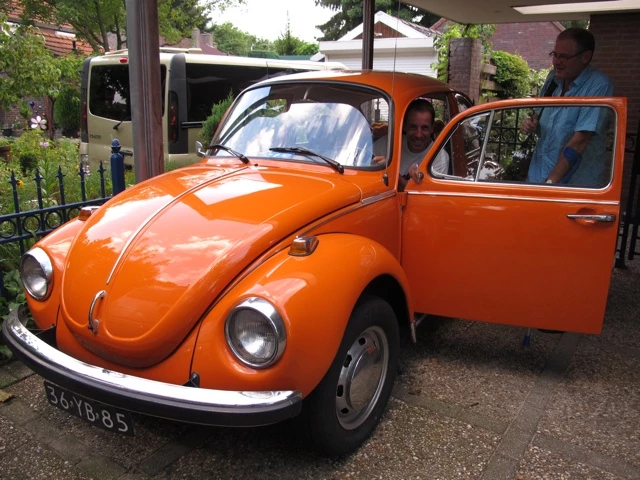 Leuchtend orangefarbener Volkswagen Käfer mit geöffneter Fahrertür, parkend vor einer Hecke und einem Gebäude
