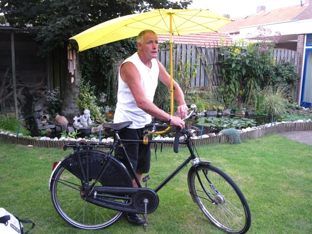 Schwarzes Fahrrad mit gelbem Sonnenschirm in einer grünen Gartenanlage, Besitzer trägt weißes Trägershirt