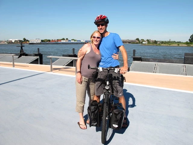 Zwei Radfahrer stehen mit einem beladenen Fahrrad auf einer Anlegestelle, Fluss und Landschaft im Hintergrund