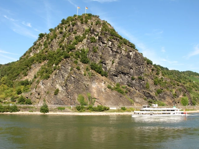 Steiler Schieferfelsen mit Vegetation am Ufer des Rheins, weißes Ausflugsschiff im Vordergrund, blauer Himmel