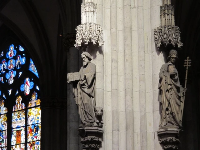 Zwei mittelalterliche Steinstatuen in gotischer Architektur, links Buntglasfenster mit blauem Lichteinfall