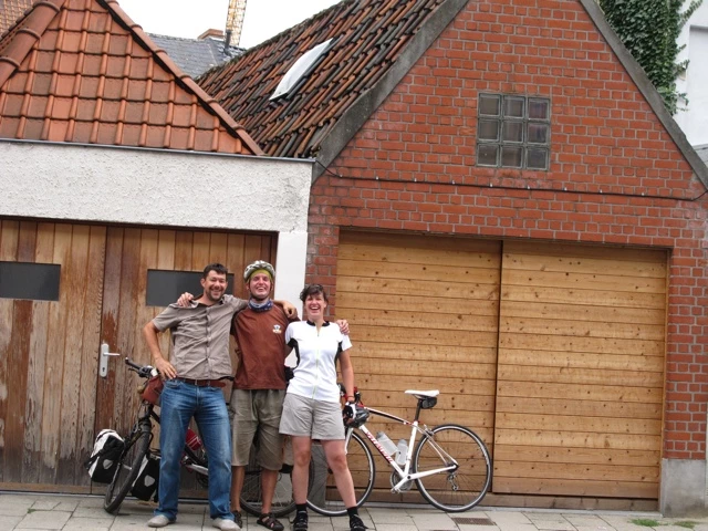 Drei Personen mit Fahrrädern stehen vor einem Backsteinhaus mit Holztor und verschiedenen Dachziegeln