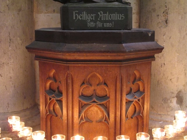 Holzständer mit gotischen Verzierungen, darauf Kerzen und Steinsockel mit deutscher Inschrift 'Heiliger Antonius'