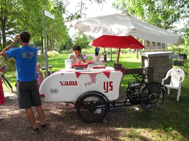 Weißer Fahrradanhänger mit Verkaufsstand, rotem Sonnenschirm und Zierbanner, auf Kiesfläche in grüner Parkanlage