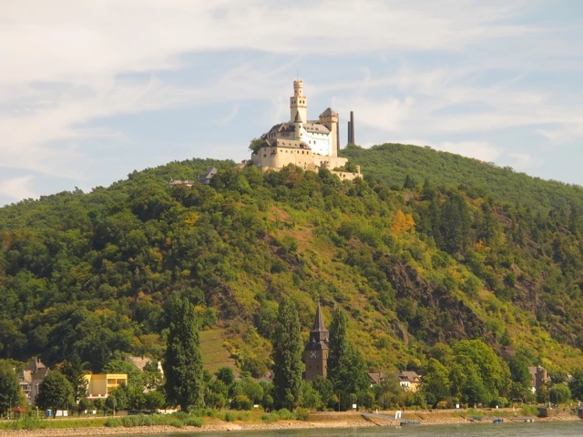 Historische Burg auf einem bewaldeten Hügel über einem Fluss, mit Stadtkulisse im Vordergrund