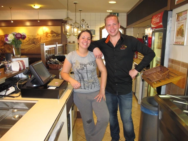 Zwei lächelnde Mitarbeiter in Arbeitskleidung hinter Theke eines Cafés, Kassensystem und Küchenbereich im Hintergrund