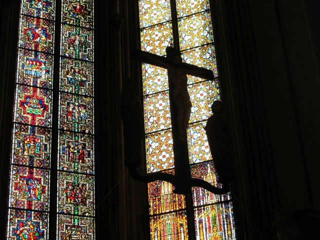 Buntes Glasfenster mit Bleiverglasung, Silhouette eines Kreuzes im Gegenlicht, links detaillierte Fenstermosaiken
