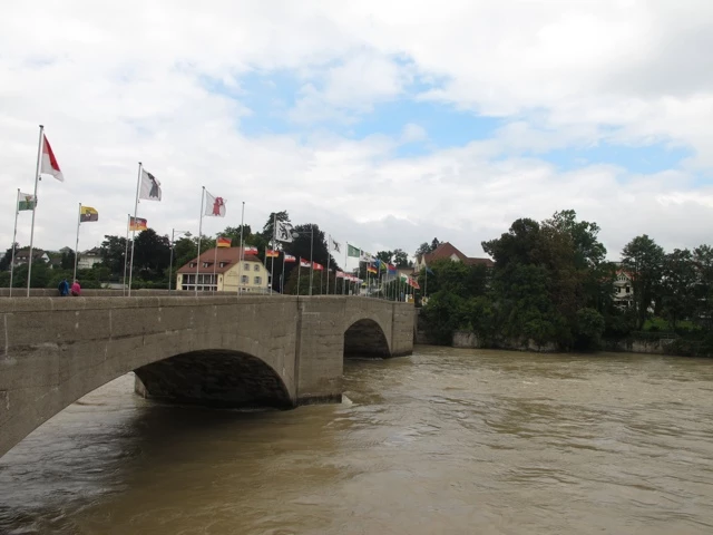 Betonbrücke mit mehreren wehenden Flaggen über einem braunen, schlammigen Fluss, Bäume und Häuser im Hintergrund