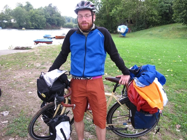 Radfahrer in blau-schwarzer Jacke und Helm steht neben Fahrrad mit Packtaschen, im Hintergrund See und Wiese