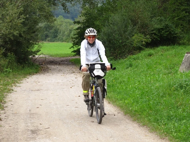 Radfahrer mit weißem Helm und Jacke fährt auf schmalem Schotterweg zwischen Wiesen und Büschen