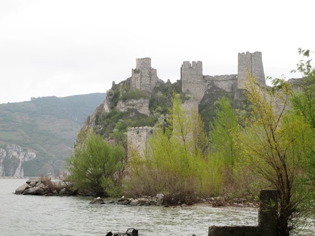 Graue Steinmauern einer historischen Burganlage auf felsigem Hang über einem Fluss mit Uferbäumen und Steinen