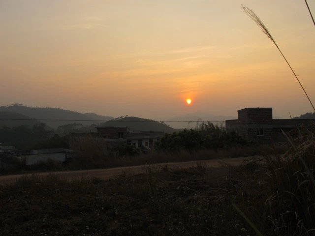 Morgendliche Landschaft mit Sonnenaufgang, Berghang, Gebäuden und Gräsern im Vordergrund in gedämpften Gelb- und Orangetönen