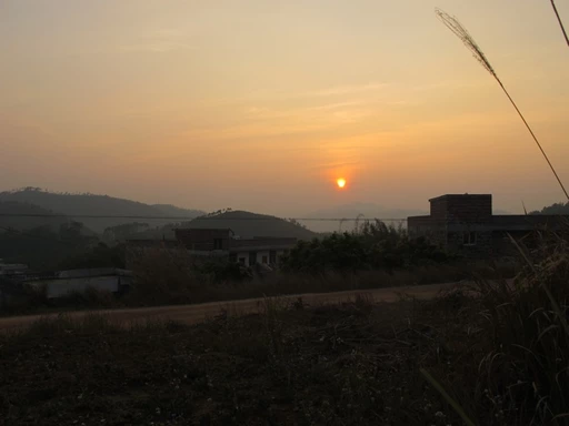 Morgendliche Landschaft mit Sonnenaufgang, Berghang, Gebäuden und Gräsern im Vordergrund in gedämpften Gelb- und Orangetönen