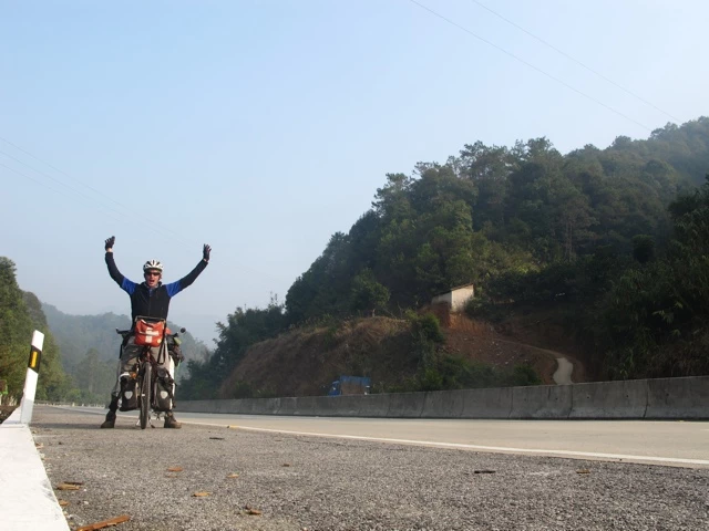 Motorradfahrer mit erhobenen Armen auf einer kurvenreichen Bergstraße in bewaldeter Landschaft, klarer Himmel