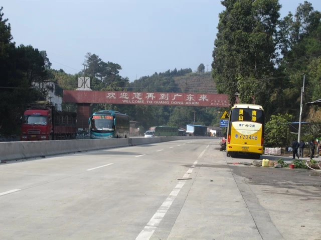 Breite Straße mit rotem Lastwagen, blauem Bus und gelbem Kleinbus, Willkommensschild im Hintergrund, bewaldete Hügellandschaft