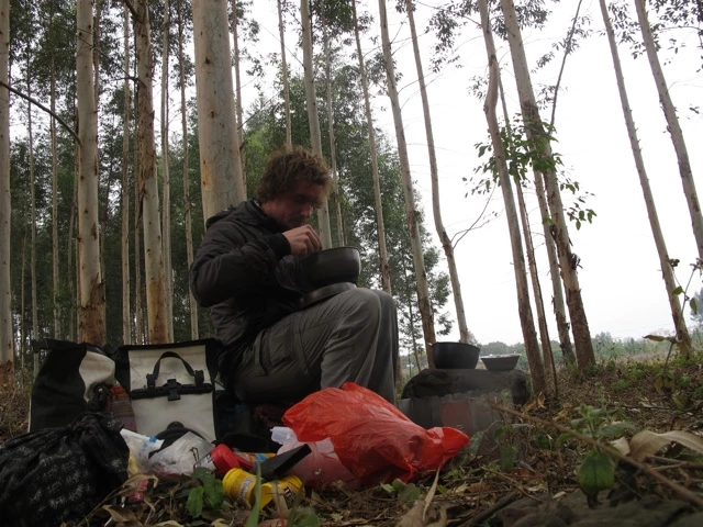 Person in dunkler Jacke sitzt auf Waldboden, umgeben von hohen Bäumen und Campingausrüstung
