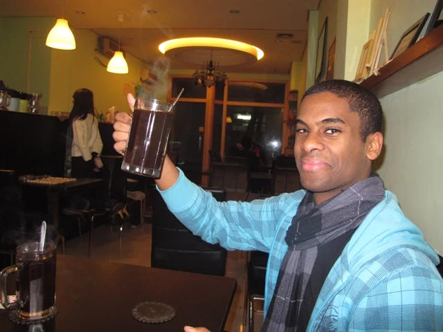 Person in blau-karierter Jacke hebt Kaffeebecher in warmbeleuchteter Caféteria