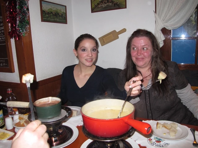 Zwei Frauen sitzen an einem Tisch mit rotem Fondue-Topf, Käse und Zutaten, lächeln und genießen gemeinsam
