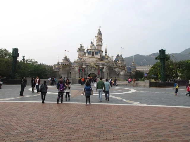 Disney-Schloss mit Eingangsportal, Menschen laufen über Pflasterstein-Vorplatz, Berge im Hintergrund
