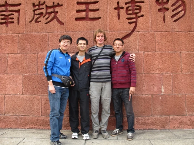 Vier junge Männer stehen eng beieinander vor einer roten Backsteinmauer mit chinesischen Schriftzeichen