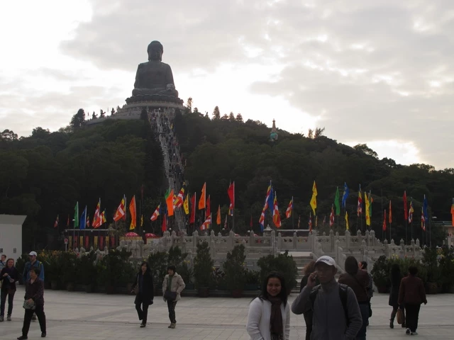 Große schwarze Buddha-Statue auf einem grünen Hügel, umgeben von bunten Flaggen und Besuchern auf einer Plattform