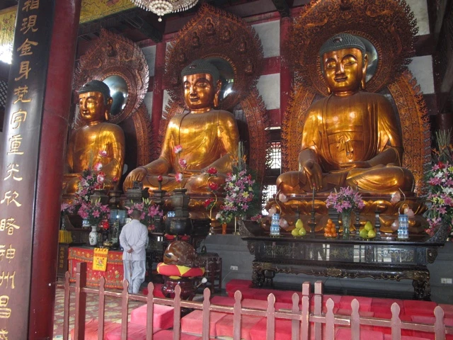 Drei große goldene Buddha-Statuen in einem buddhistischen Tempel, davor Blumen und eine Person in weißer Kleidung