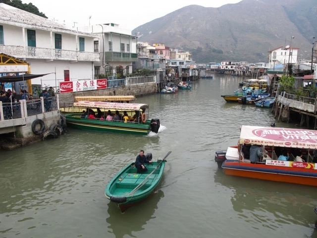 Grüne und bunte Boote schwimmen in einem schmalen Wasserkanal mit Häusern, Bergen im Hintergrund und Coca-Cola-Werbung