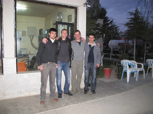 Vier junge Männer stehen zusammen vor einem Gebäude, dahinter weiße Stühle und Bäume in der Abenddämmerung