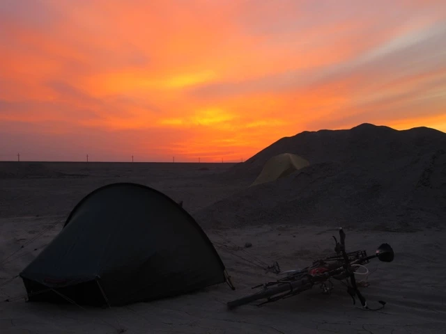 Schwarzes Zelt und liegendes Fahrrad vor dramatischem orangefarbenen Sonnenuntergangshimmel und Bergsilhouette in karger Wüstenlandschaft