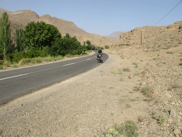 Schmale Asphaltstraße durch trockene Berglandschaft mit Motorrad, grünen Bäumen links und felsiger Wüstenlandschaft rechts
