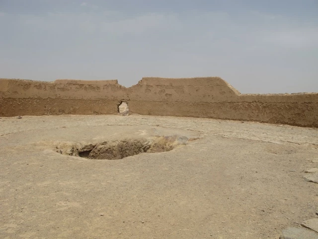 Trockene Lehmwände einer alten Siedlung mit einem Erdloch im Vordergrund, flache Wüstenebene mit blassem Himmel