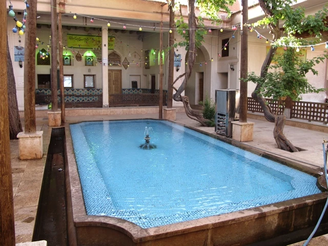 Blauer rechteckiger Pool in überdachtem Innenhof mit Holzstützen, Bäumen und Mosaikfliesen