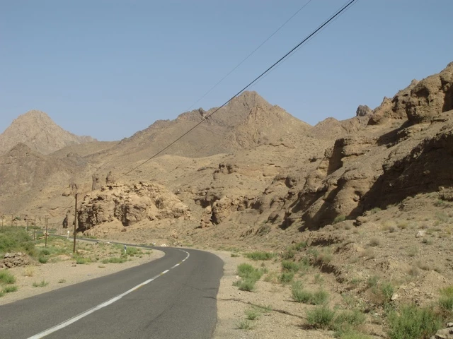 Gewundene Asphaltstraße führt durch felsige, braune Berglandschaft mit karger Vegetation und Stromleitungen