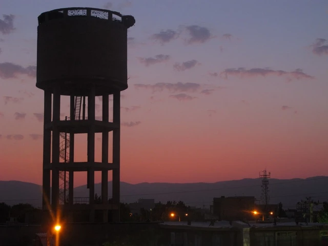 Schwarzer Wasserturm mit Stahlkonstruktion vor Bergsilhouette, Abendhimmel in Orange und Rosa