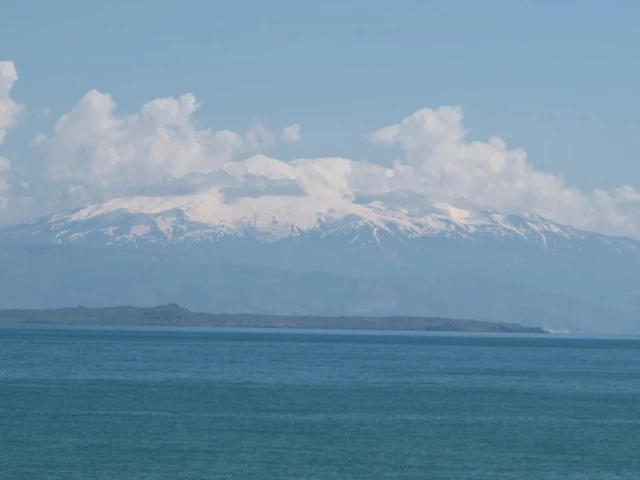 Weite Landschaft mit schneebedeckten Berggipfeln, die sich über einem türkisblauen See erheben, vor blauem Himmel mit Wolken
