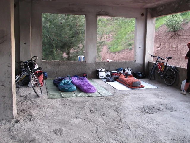 Fahrräder und Schlafsäcke in einer unfertigten Betonhalle mit Ausblick auf grüne Hügellandschaft
