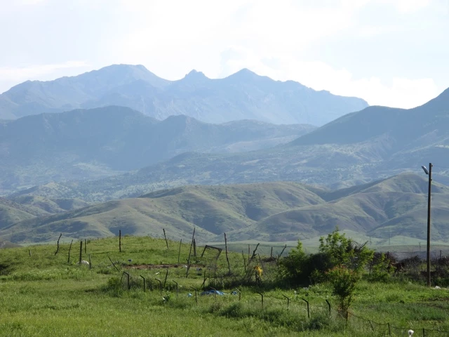 Weite Berglandschaft mit sanften Hügeln, grüner Wiese im Vordergrund und blau-grauen Berggipfeln im Hintergrund