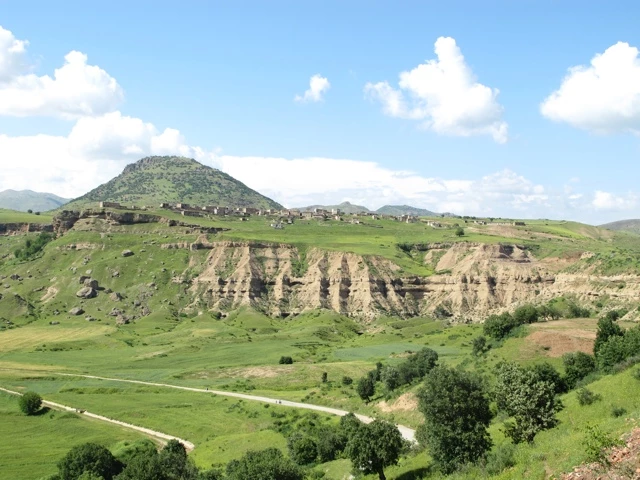 Grüne Hügellandschaft mit Erosionsrinnen, Dorf auf Bergkuppe, serpentineartige Straße durch saftig-grüne Wiesen