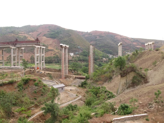 Große Betonbrückenpfeiler im Bau, eingebettet in hügelige Landschaft mit Straßen und spärlicher Vegetation