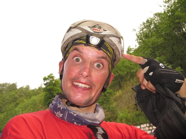 Nahaufnahme eines Radfahrers mit Helm, roter Jacke und Handschuhen, der einen übertrieben überraschten Gesichtsausdruck macht, grüne Vegetation im Hintergrund