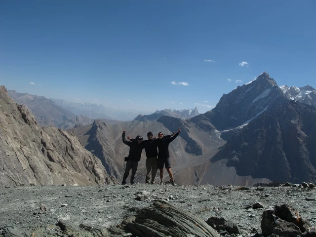 Drei Wanderer stehen vor imposanter Bergkulisse mit Schneebergen, steiniger Vordergrund, klarer blauer Himmel
