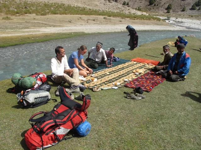 Reisegruppe sitzt an einem Fluss auf Decken, umgeben von Rucksäcken und Ausrüstung, Berglandschaft im Hintergrund