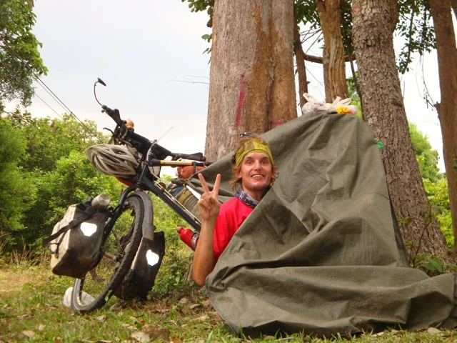 Fahrrad mit Packtaschen steht neben grünem Zelt zwischen Bäumen, Radfahrer trägt gelbe Mütze und macht Friedenszeichen